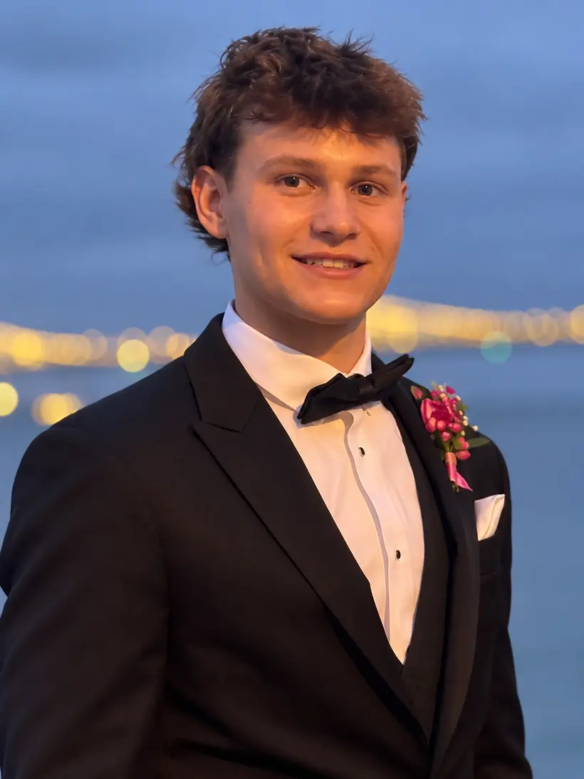 Shea smiling in a black tuxedo with a pink boutonniere, in front of city lights over the water at twilight
